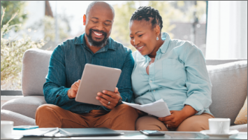 Man and woman sitting on couch. Woman is holding papers and man is holding a tablet. Both are looking at the tablet.