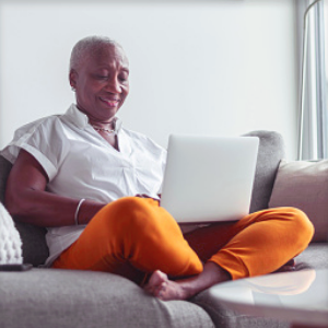 A woman sits cross-legged on a couch with a laptop on her lap.