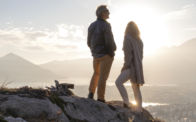 Man and woman in silhouette standing on rocks overlooking a lake and mountains at sunset