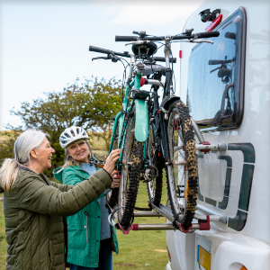 Two retired women put bicycles on a rack on the back of a motor coach.