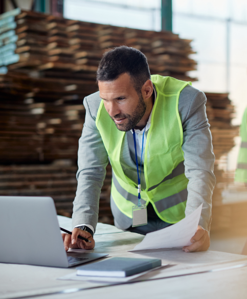 Man wearing neon vest over suit looks at laptop in warehouse