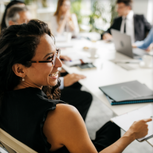 A woman sits at a conference table and smiles with others out of focus in the background.