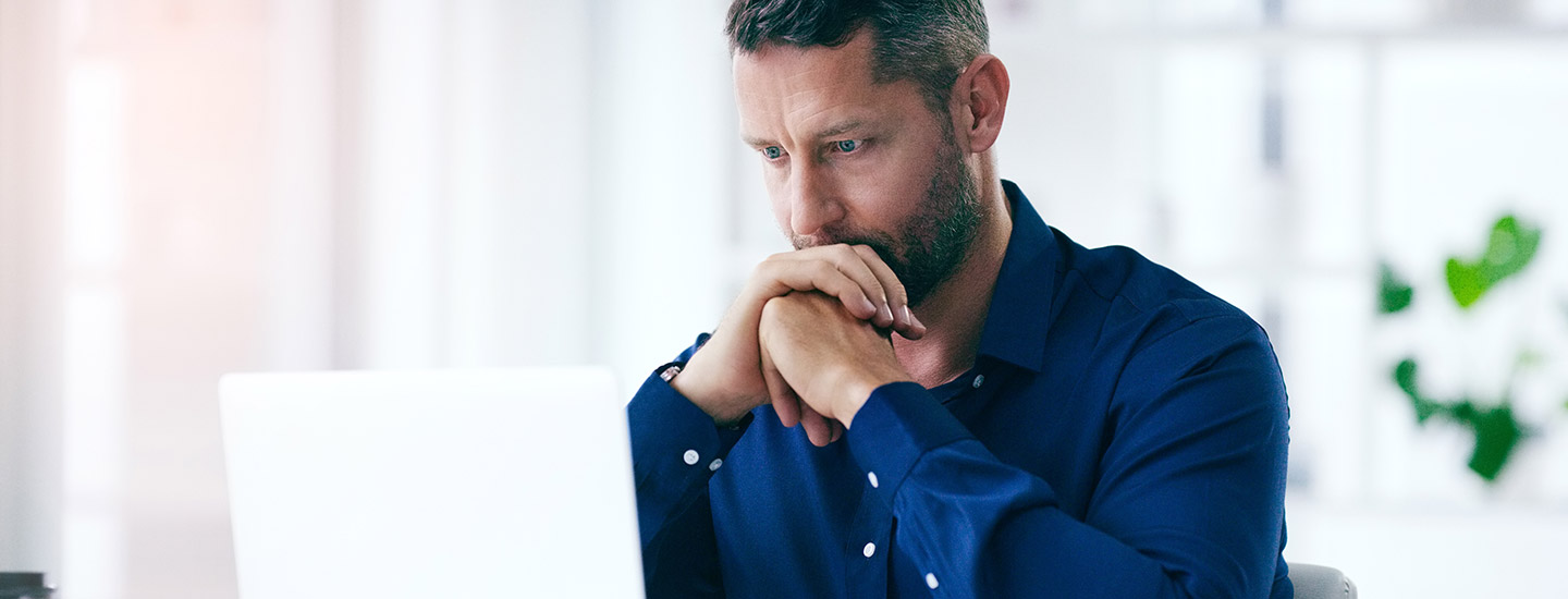 Man with hands together against his mouth looks at laptop with furrowed brows