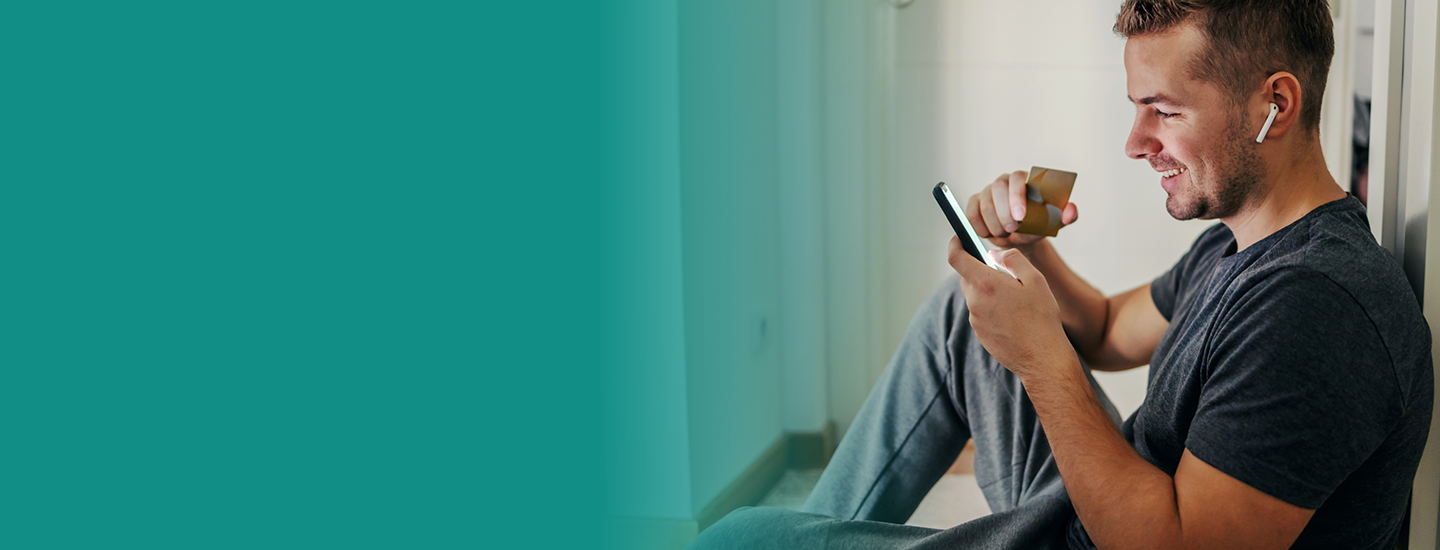 A man sits on the floor holding a phone and a credit card and wearing headphones.