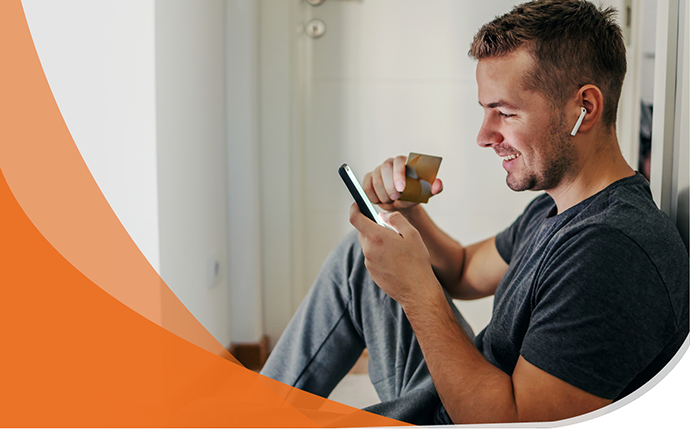 A man sitting on the floor looking at his phone, holding a credit card, and wearing headphones.