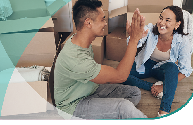 A young couple high fives on the floor of a kitchen surrounded by moving boxes.