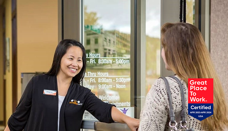 A female IncredibleBank employee holds open the door for a woman entering the branch.